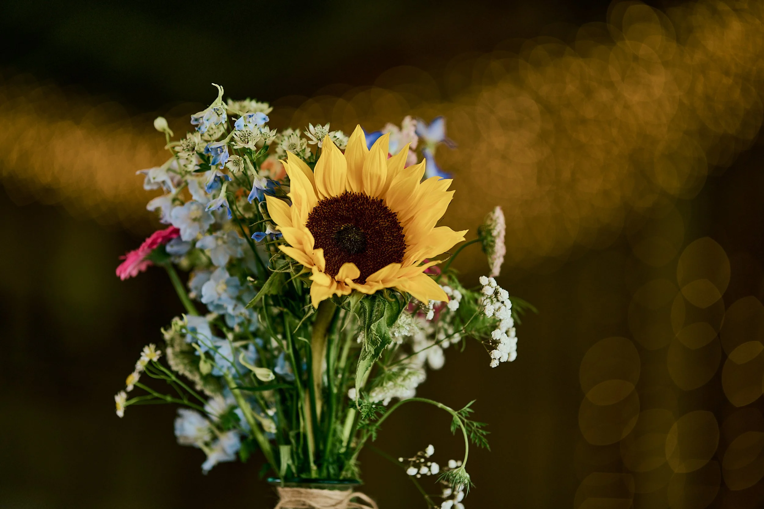 A bouquet of mixed flowers including a prominent yellow sunflower against a blurred dark background with bokeh light effects.