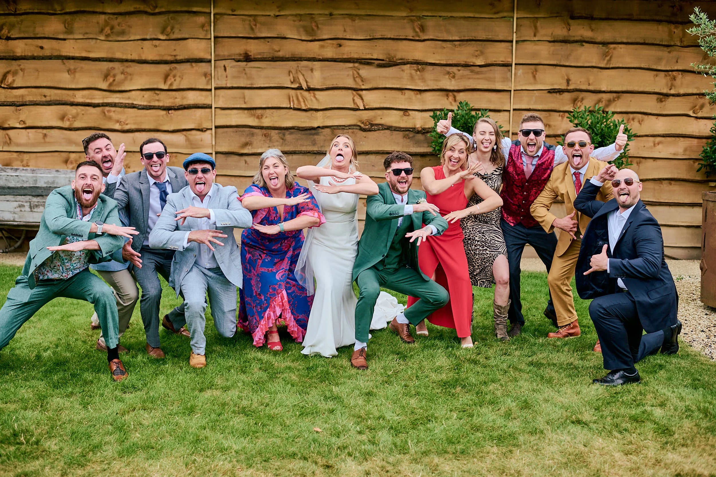 A group of 14 people dressed in colorful and formal clothing, wearing sunglasses, standing on grass in front of a wooden wall, making funny faces and poses, celebrating at an outdoor event.