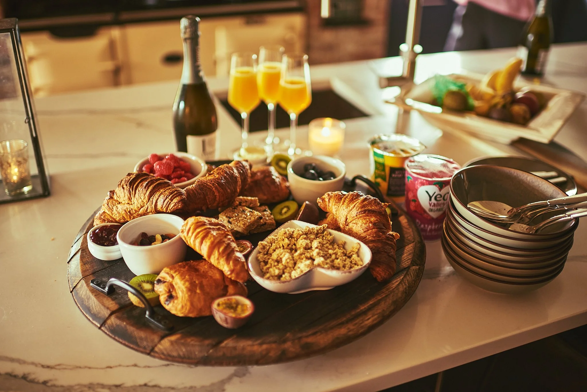 A breakfast spread on a round wooden platter includes croissants, raspberries, blackberries, and sliced kiwi, along with small bowls of jam, yogurt, and crumble. In the background, there are three glasses of orange juice, a bottle of champagne, and a