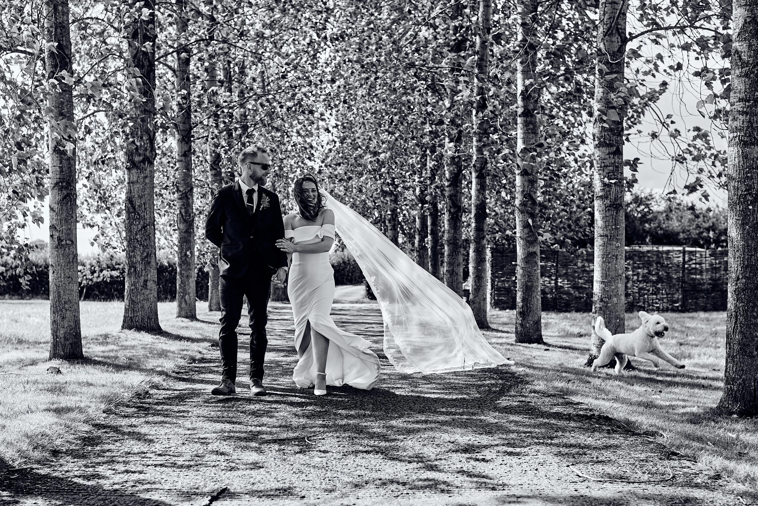 Black and white photo of a bride and groom walking along a tree-lined path outdoors, smiling, with a dog running nearby.