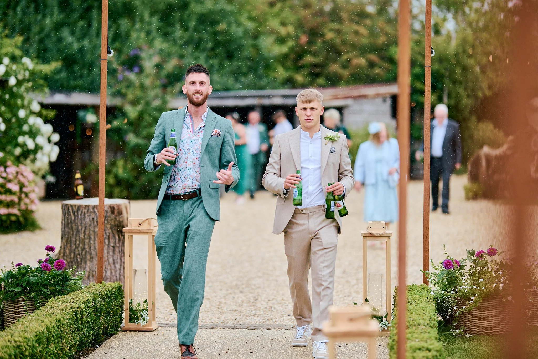 Two men in suits walking outdoors at a wedding or celebration, holding green bottles, with a string of wooden posts and flowers in the foreground.