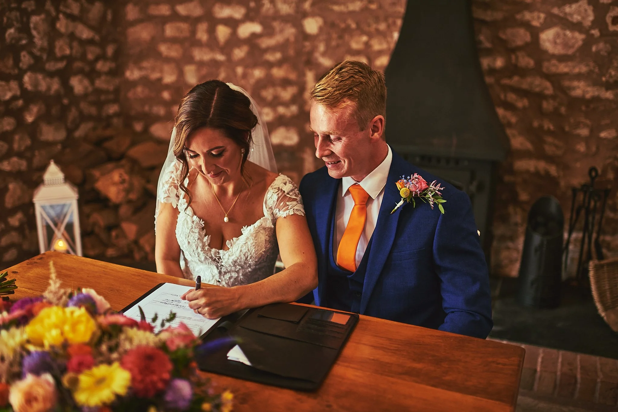 A bride and groom signing a marriage document at their wedding ceremony in a warmly lit, rustic indoor setting with a stone wall background. The bride wears a white lace dress and the groom is in a navy suit with an orange tie, both smiling.