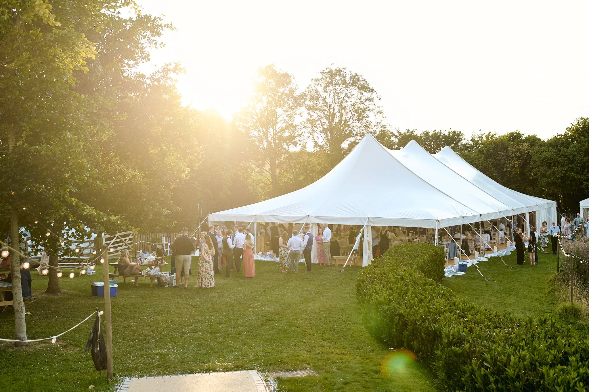 Guests gathered under a white event tent with peak tops at an outdoor party in the late afternoon, surrounded by green trees and bushes, with string lights on a nearby tree.