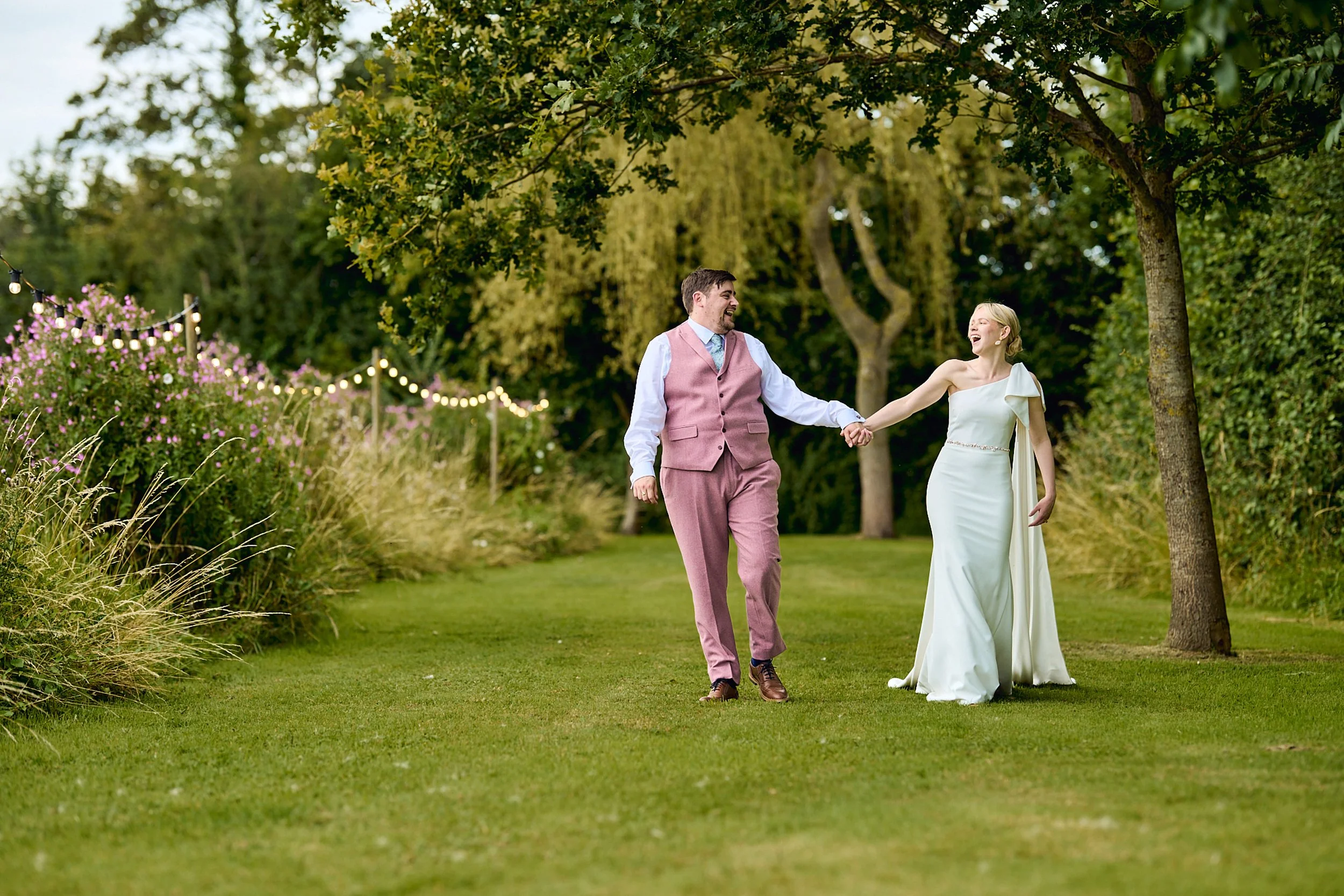 A bride and groom holding hands and smiling while walking through a lush green garden.