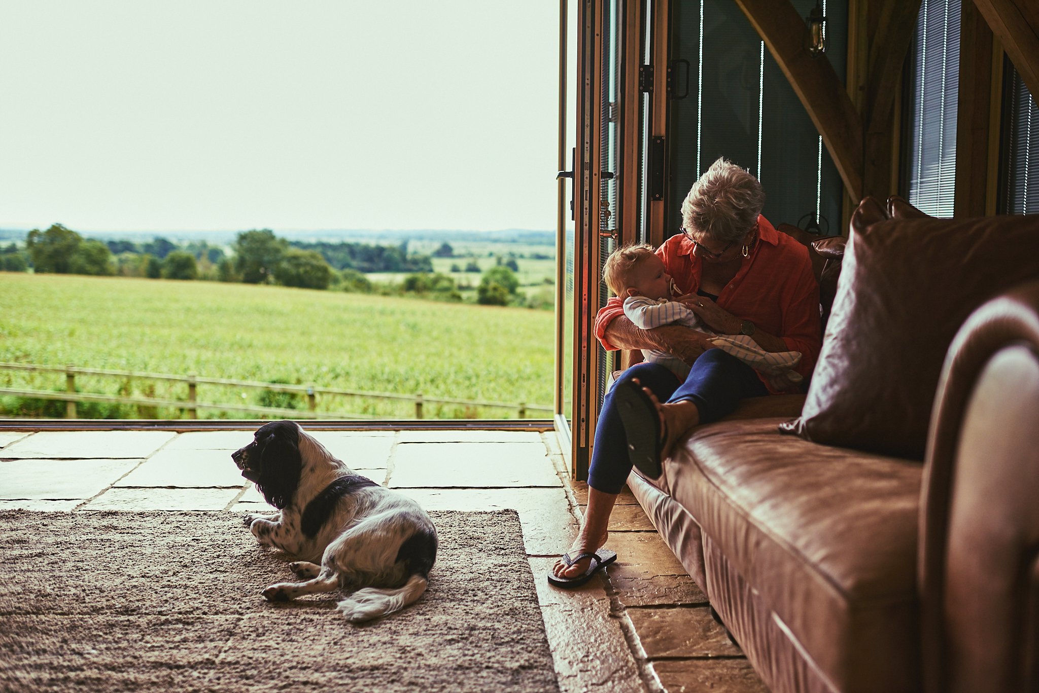 An elderly woman and a young child sitting on a couch inside a room, looking at each other affectionately, with a dog lying on a rug in front of them and an open door showing a green field and trees outside.