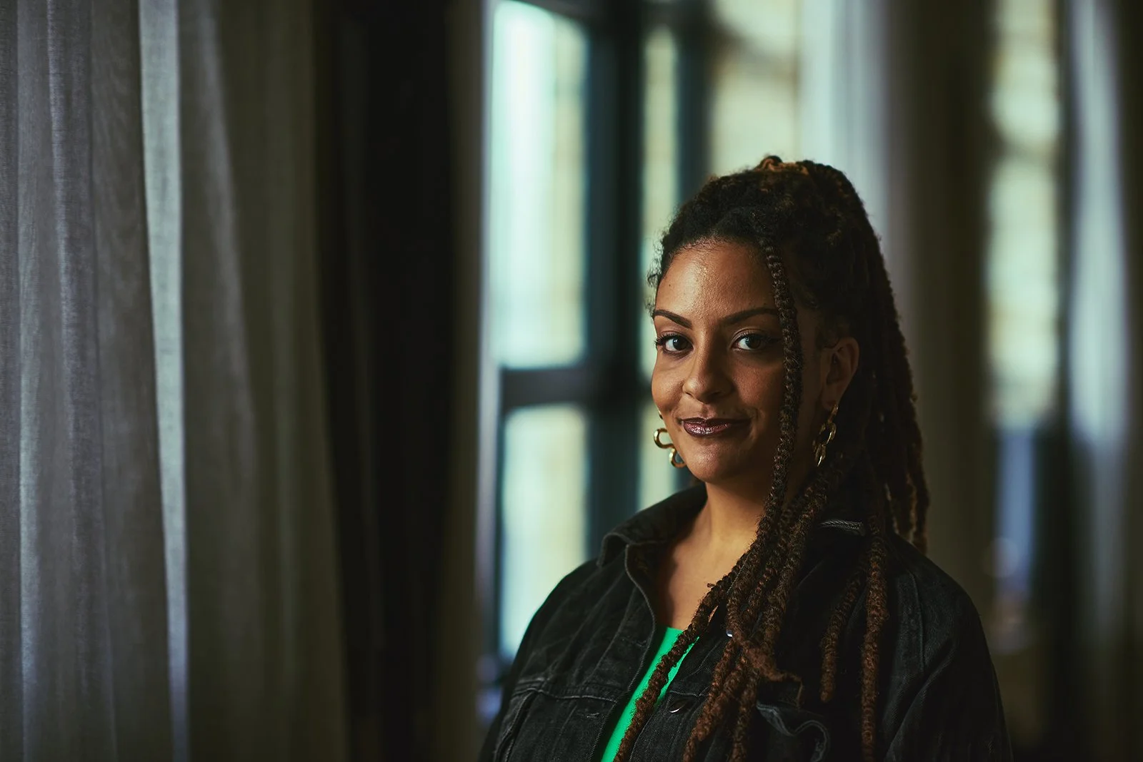 A woman with dreadlocks, wearing a dark jacket, standing by a window in a dimly lit room with curtains.