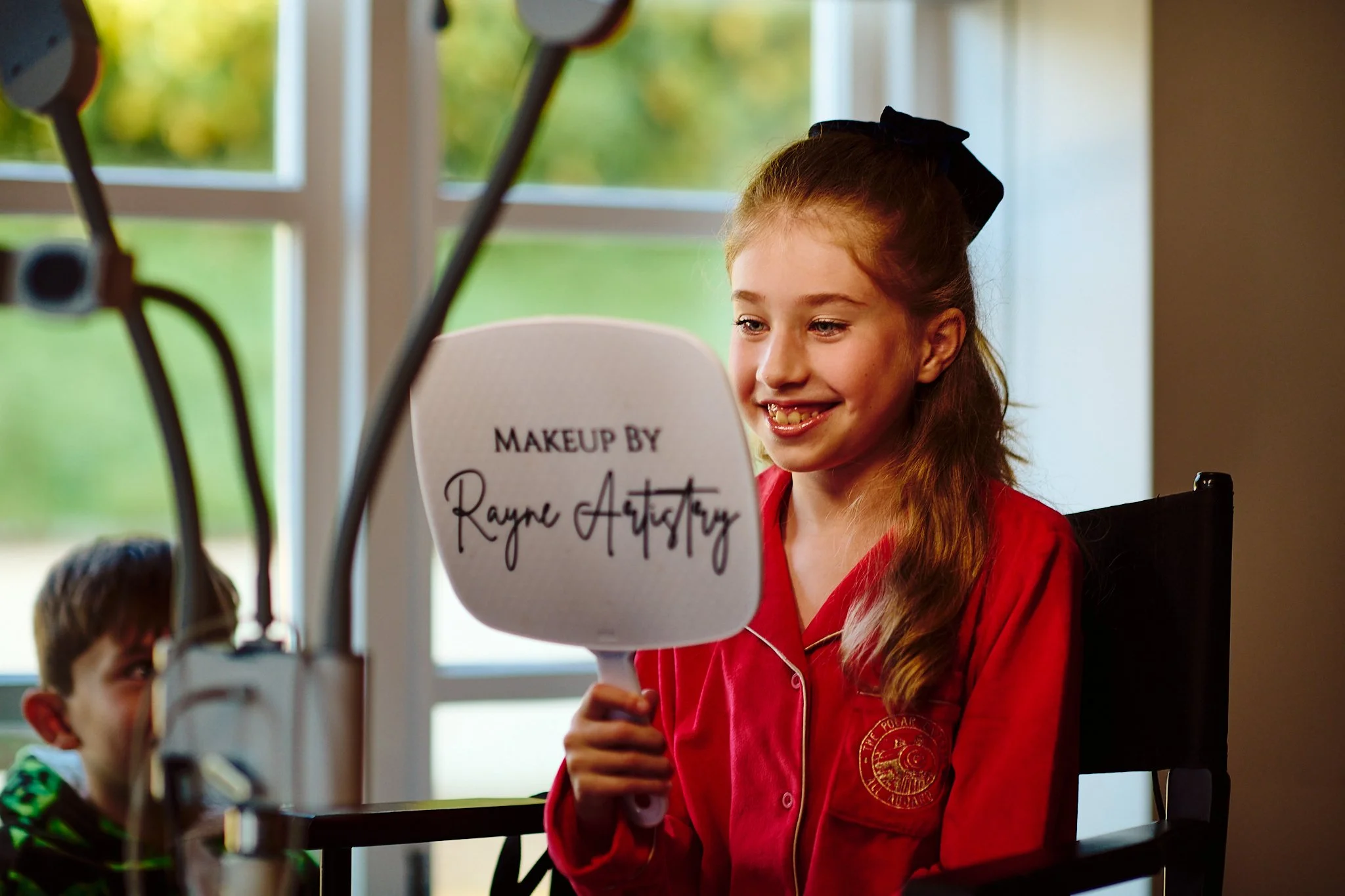 A young girl with long red hair and a black bow, wearing a red jacket, smiling and holding a makeup artist sign in front of a makeup station with a mirror and other children, near a window with green trees outside.
