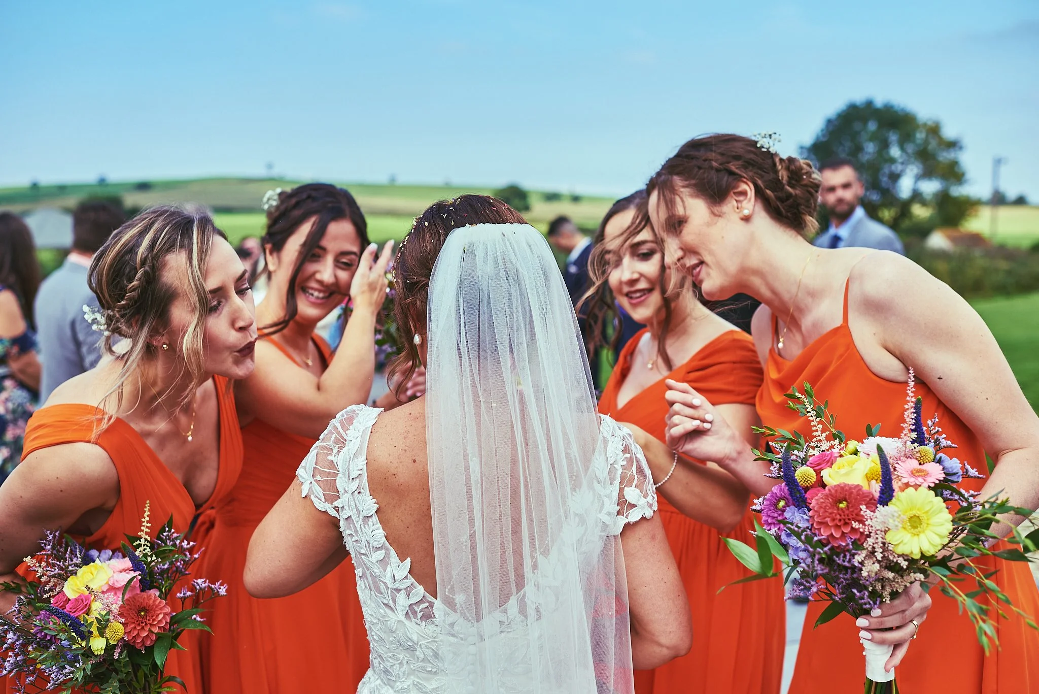 Bride in a white lace dress and veil surrounded by bridesmaids in orange dresses during a wedding celebration outdoors with a green landscape and cloudy sky in the background.