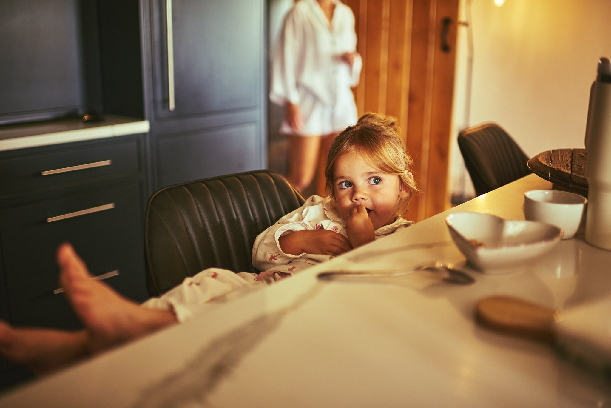 A young girl with blonde hair and blue eyes sitting at a kitchen table, looking up and slightly to the side, with her finger near her mouth. In the background, a woman is standing, partially blurred.”}