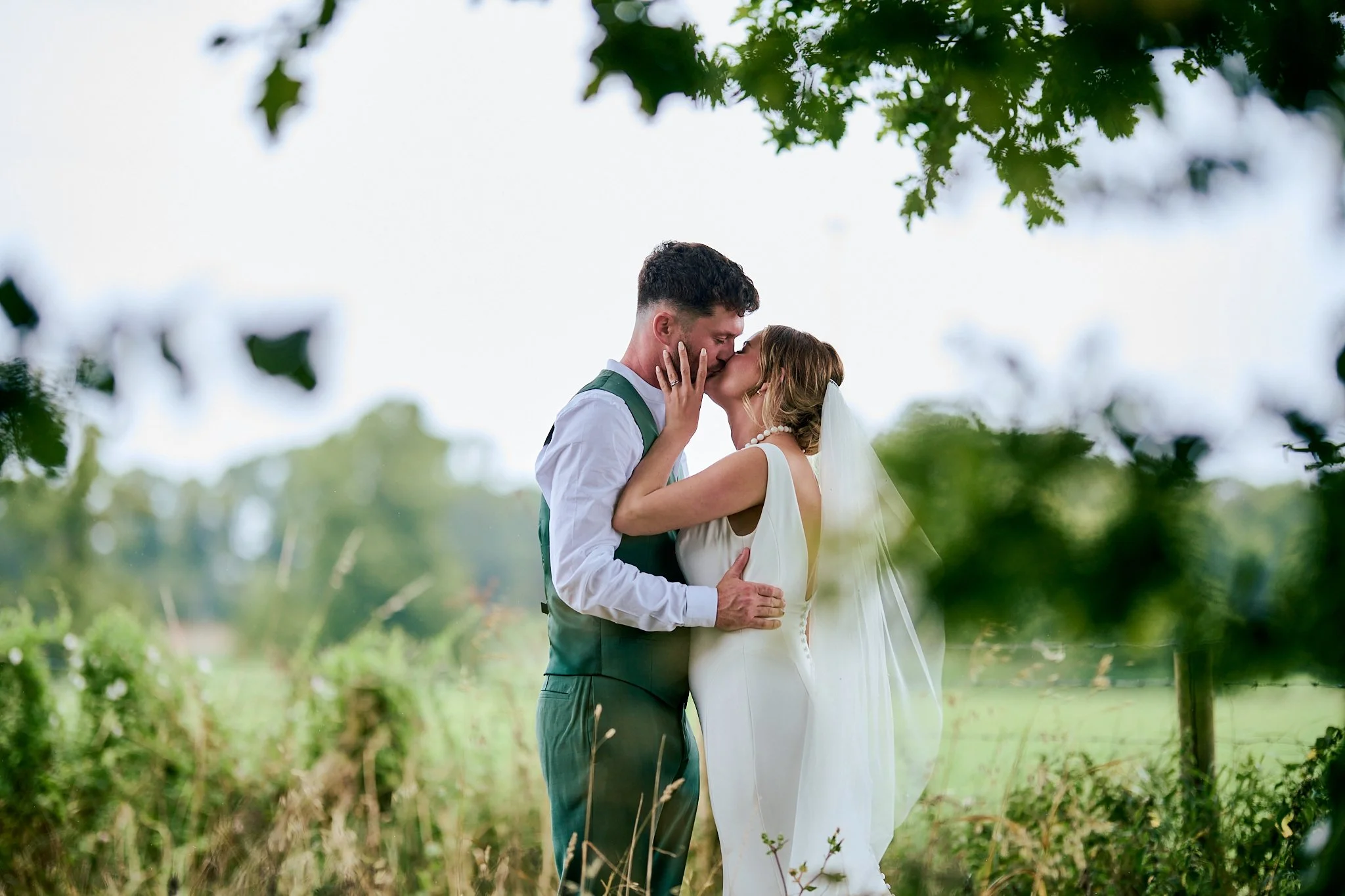 A couple sharing a kiss outdoors during their wedding, with greenery and trees in the background.