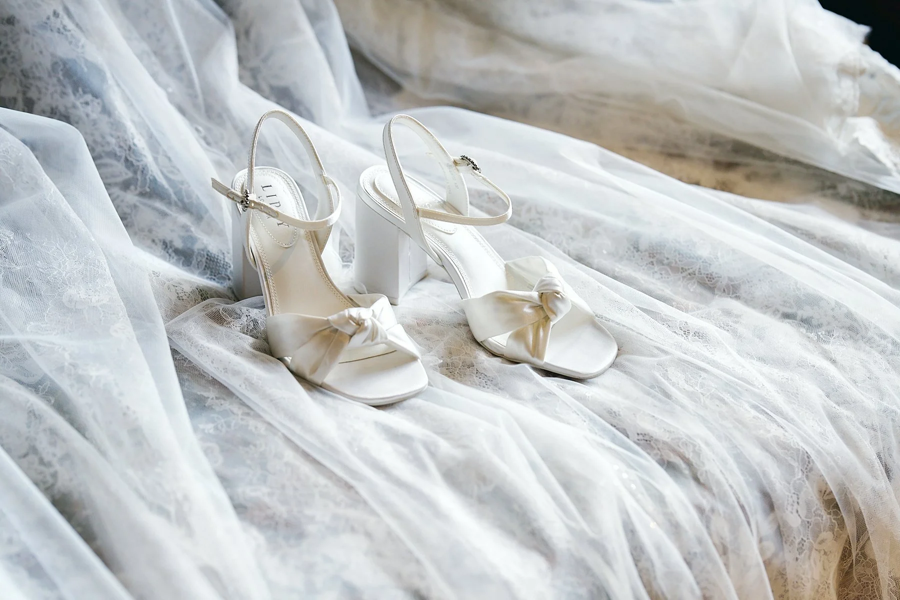 A pair of elegant white high-heeled sandals with satin bows on the toes and ankle straps, placed on lace fabric.