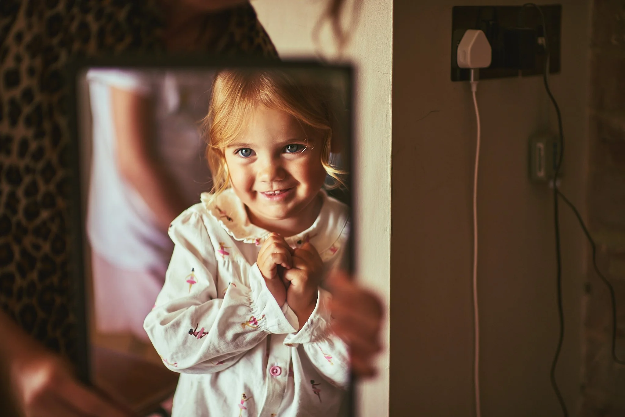 A young girl with red hair and blue eyes looking through a mirror, smiling with her hands clasped in front of her. She is wearing a white shirt with colorful designs.