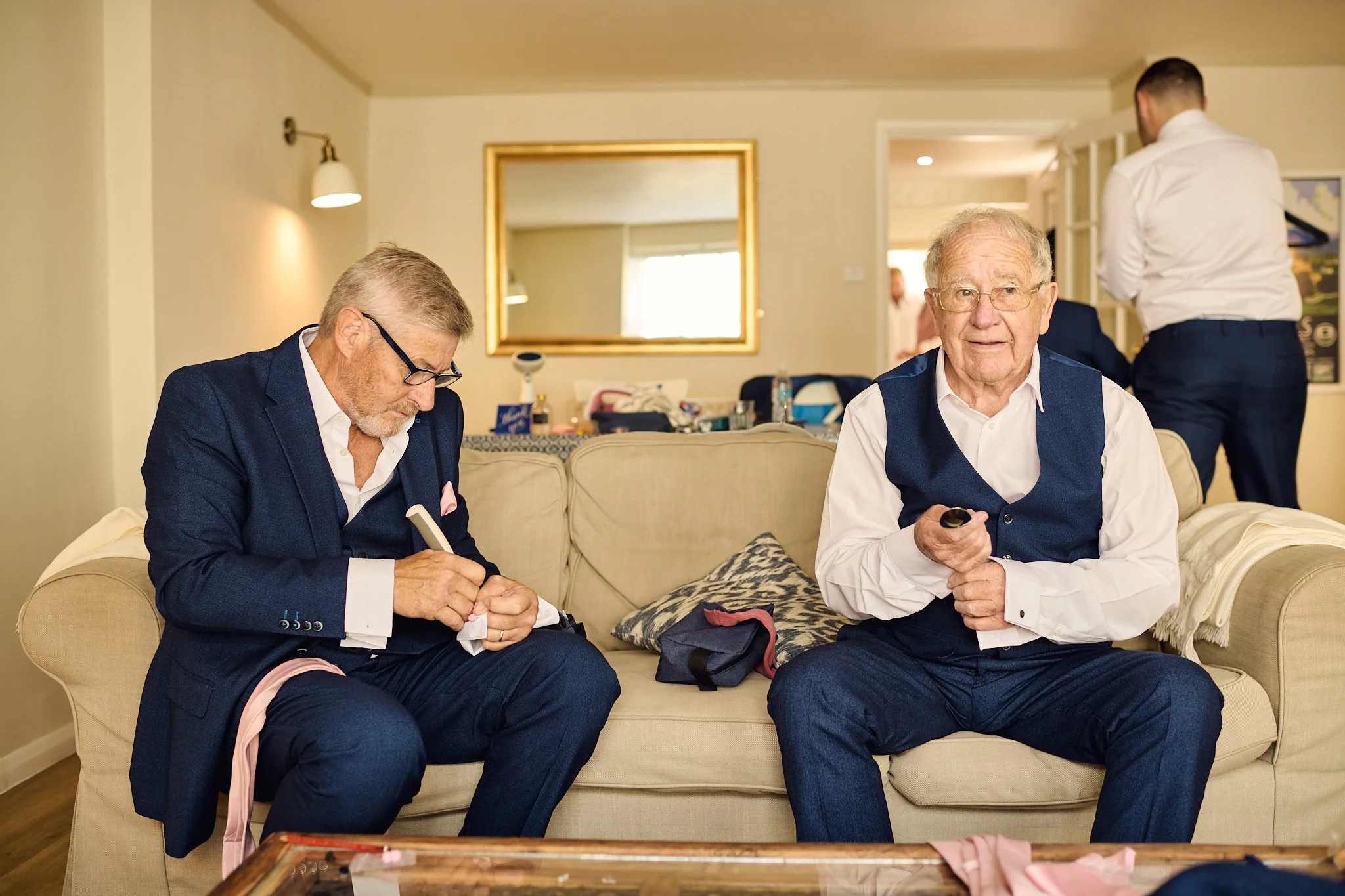 Two elderly men in formal suits sitting on a beige sofa in a living room, one fixing his cuff links and the other holding sunglasses, with a man in a white shirt and dark pants standing in the background.