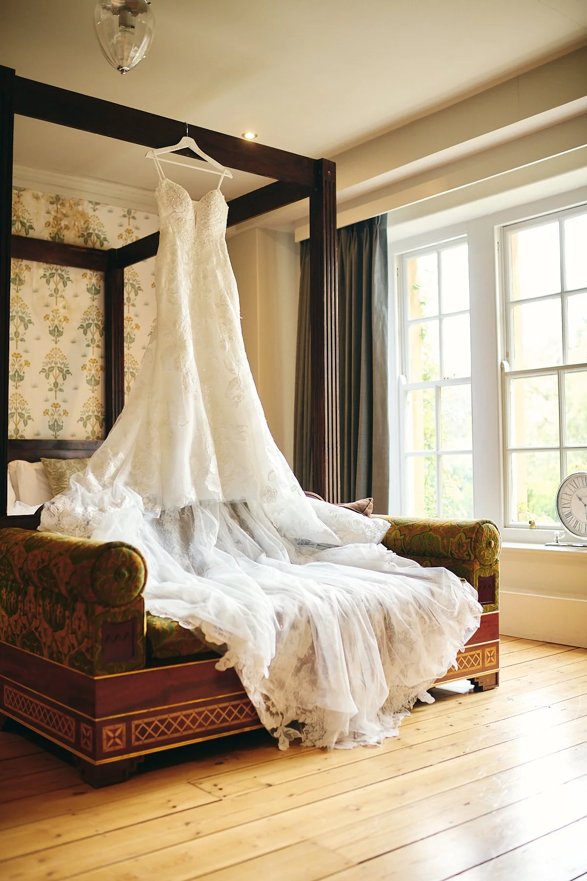 Wedding dress hanging on a four-poster bed in a sunlit room with floral wallpaper and large windows.