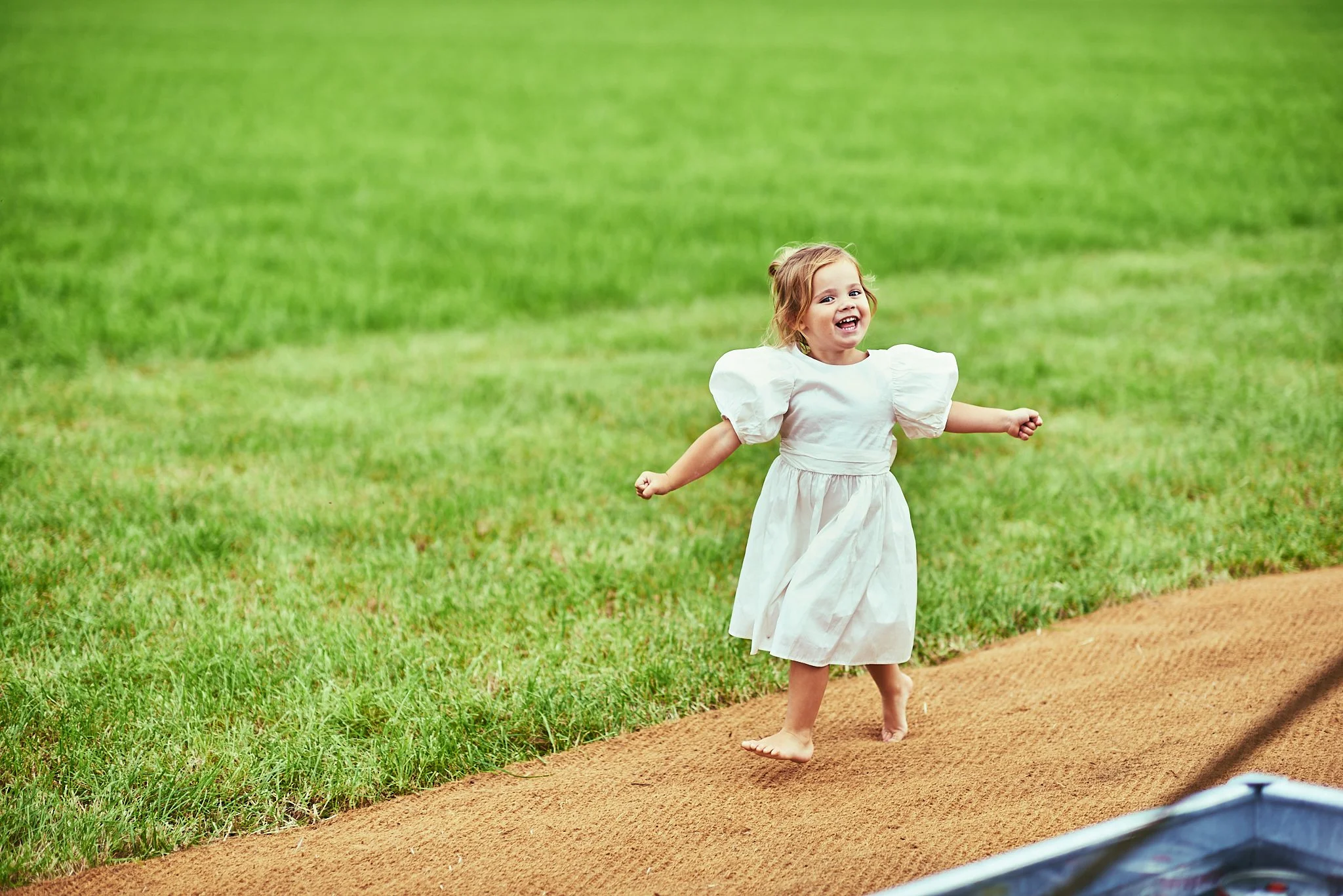 A young girl with red hair wearing a white dress running barefoot on a dirt path in a grassy field.
