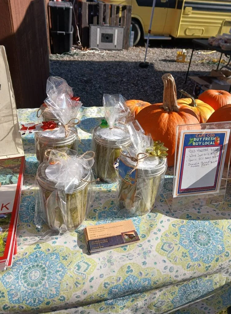 Small jars decorated with ribbons and bows, displayed on a table with pumpkins in the background, at a local market stand promoting fresh, local produce in Cape Cod.