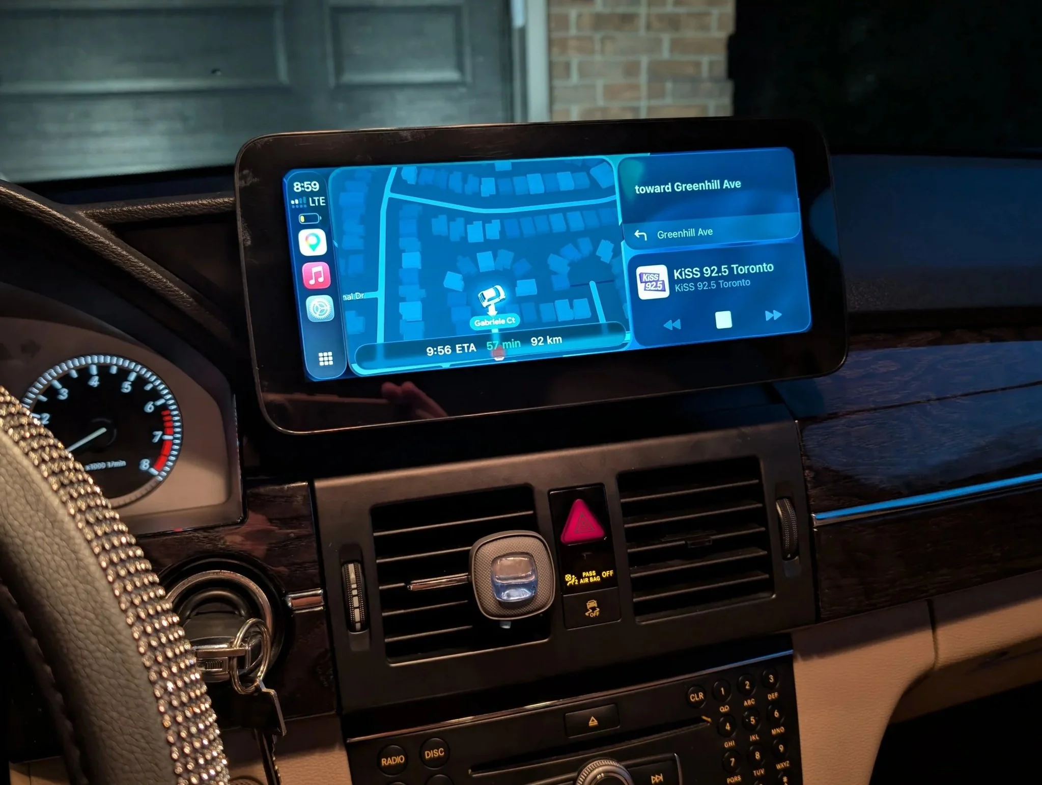 Car dashboard with a navigation screen showing a map, current time 8:59, and radio station KISS 92.5 Toronto, with the current route indicating toward Greenhill Ave and an ETA of 9:56, 57 minutes remaining, 92 km distance.