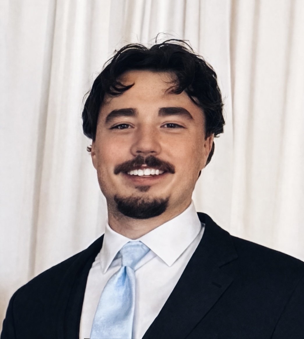 A smiling man in a formal black suit with a light blue tie and white shirt, standing in front of a light-colored curtain. Owner of Auto Advisor | Car Buying Consultant