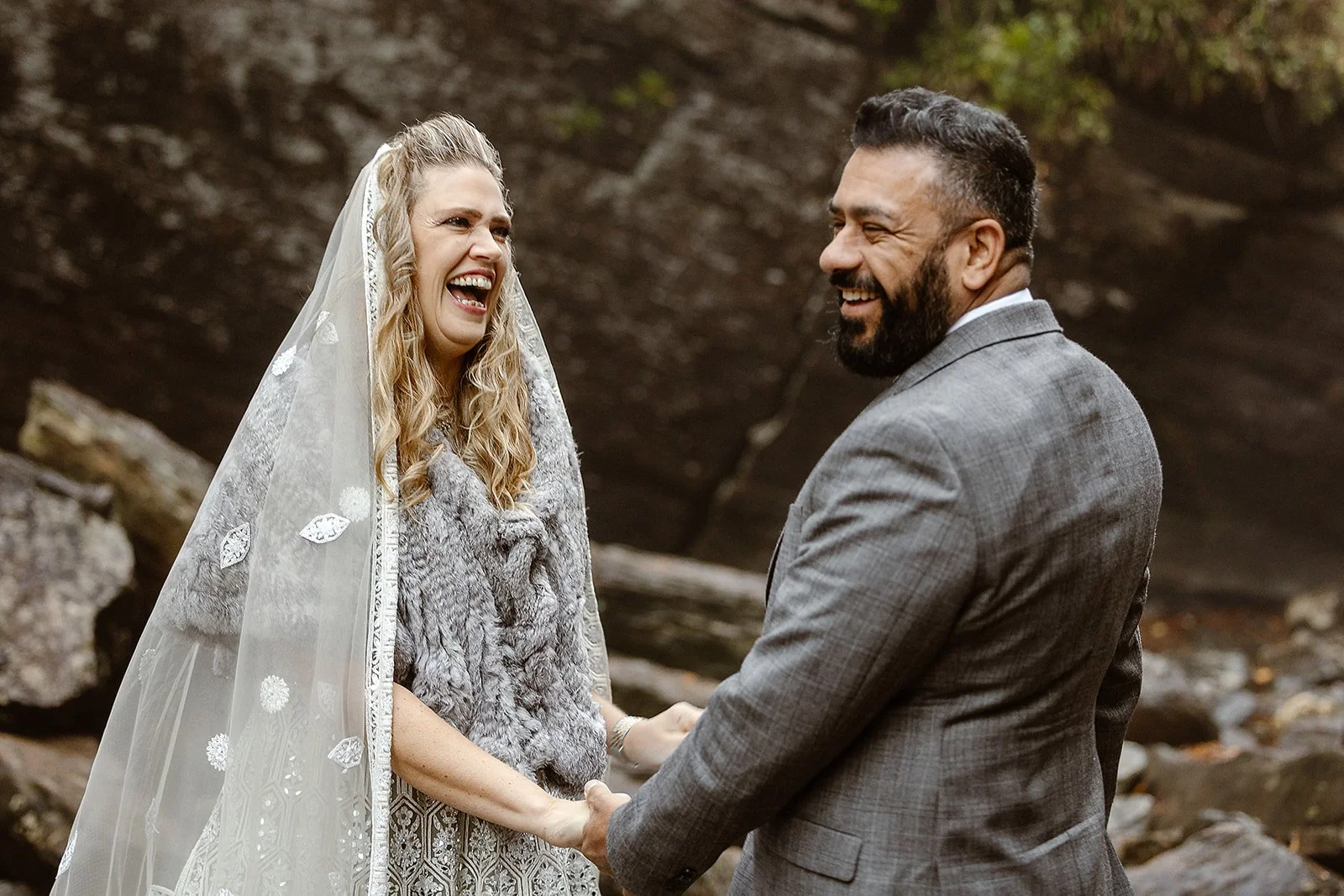A couple, dressed in wedding attire, holding hands and smiling at each other outdoors with rocks and trees in the background.