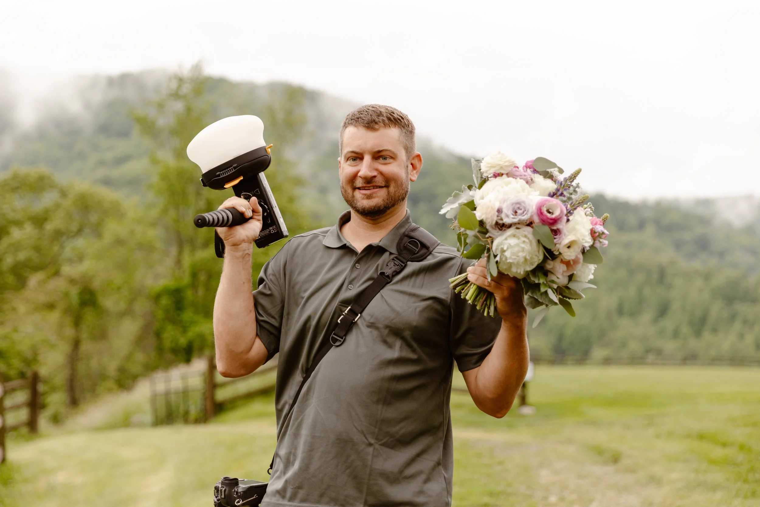 A smiling man holding a bouquet of flowers in one hand and a paintball gun in the other, standing outdoors on a grassy field with trees and a fence in the background.
