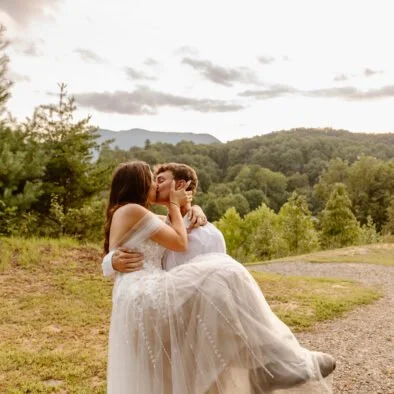 A couple in wedding attire sharing a kiss outdoors with a scenic landscape of trees and mountains in the background.