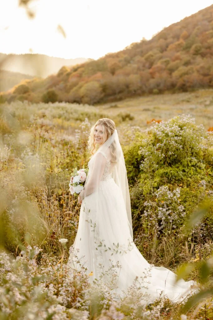 A smiling bride in a white wedding dress holding a bouquet of flowers, standing in a lush field surrounded by wildflowers and greenery with a mountain background during sunset.