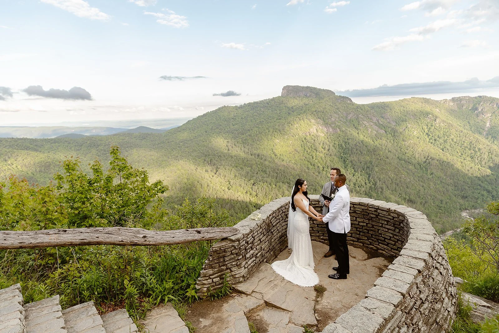 A wedding ceremony taking place on a stone platform with a scenic mountain view in the background, featuring a bride and groom holding hands facing each other with an officiant standing behind.