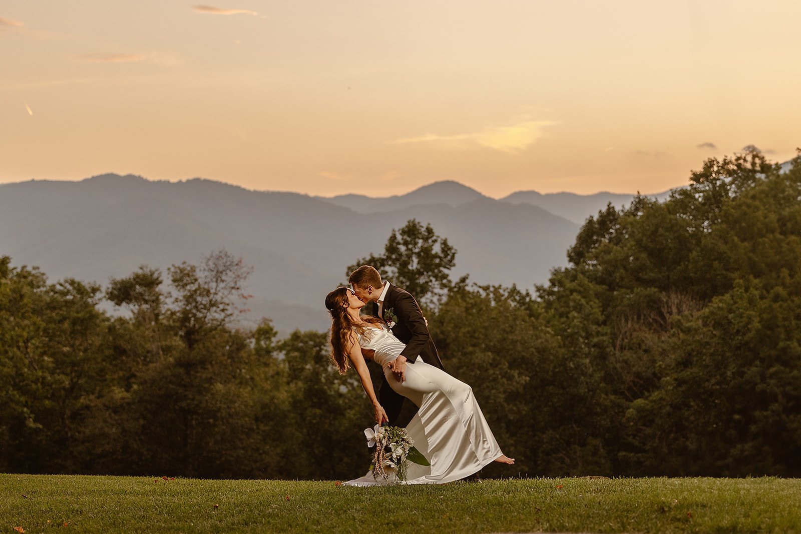 A bride and groom sharing a kiss outdoors during sunset, with mountains and trees in the background.