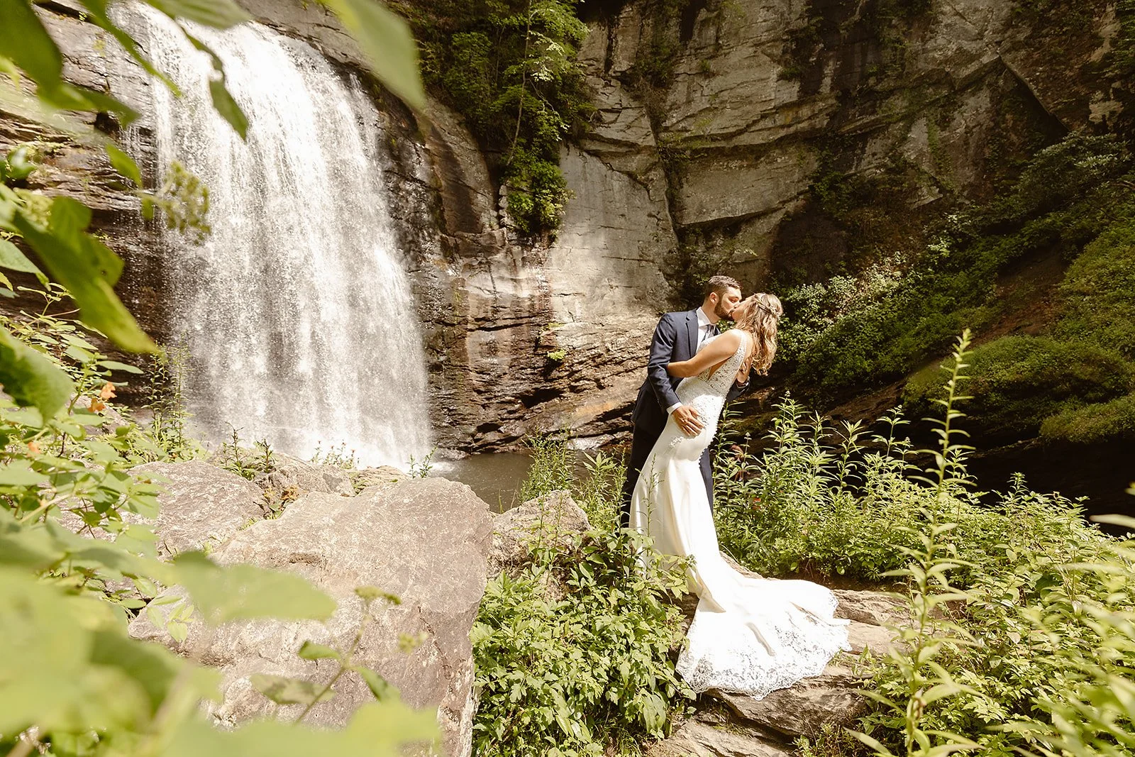 A bride and groom share a kiss in front of a waterfall with lush green foliage surrounding them.
