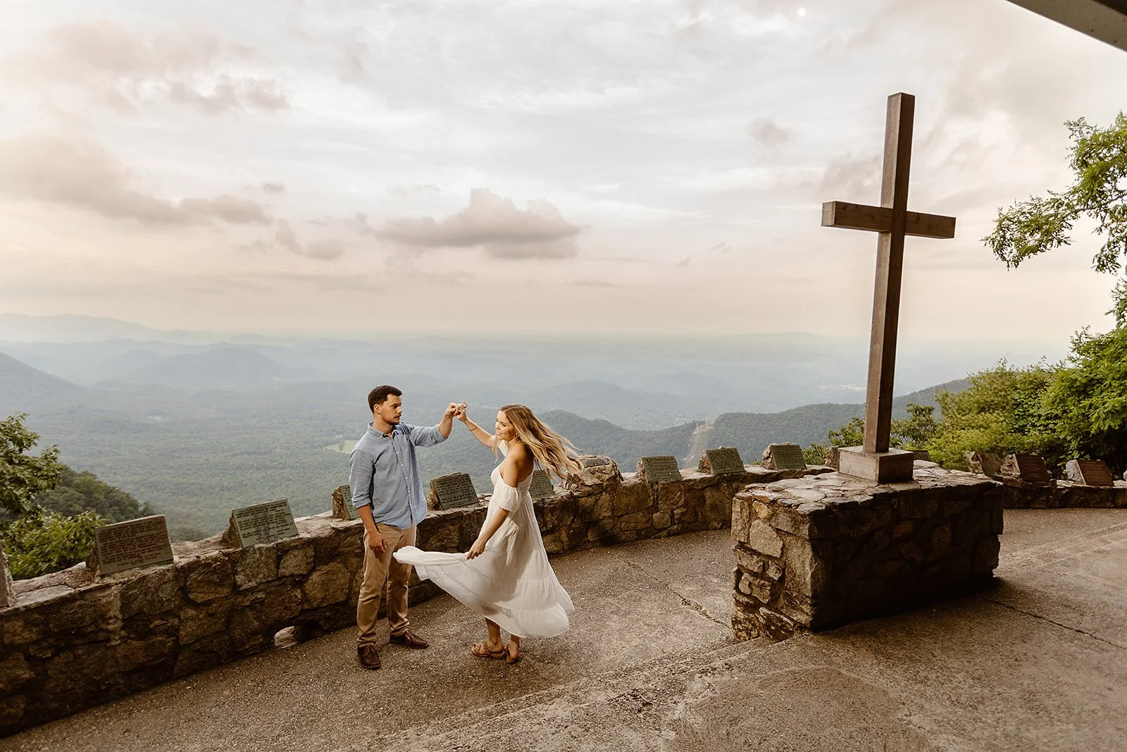 A couple dancing on a mountain overlook with a large wooden cross, overlooking a valley with hills and trees, during cloudy weather.