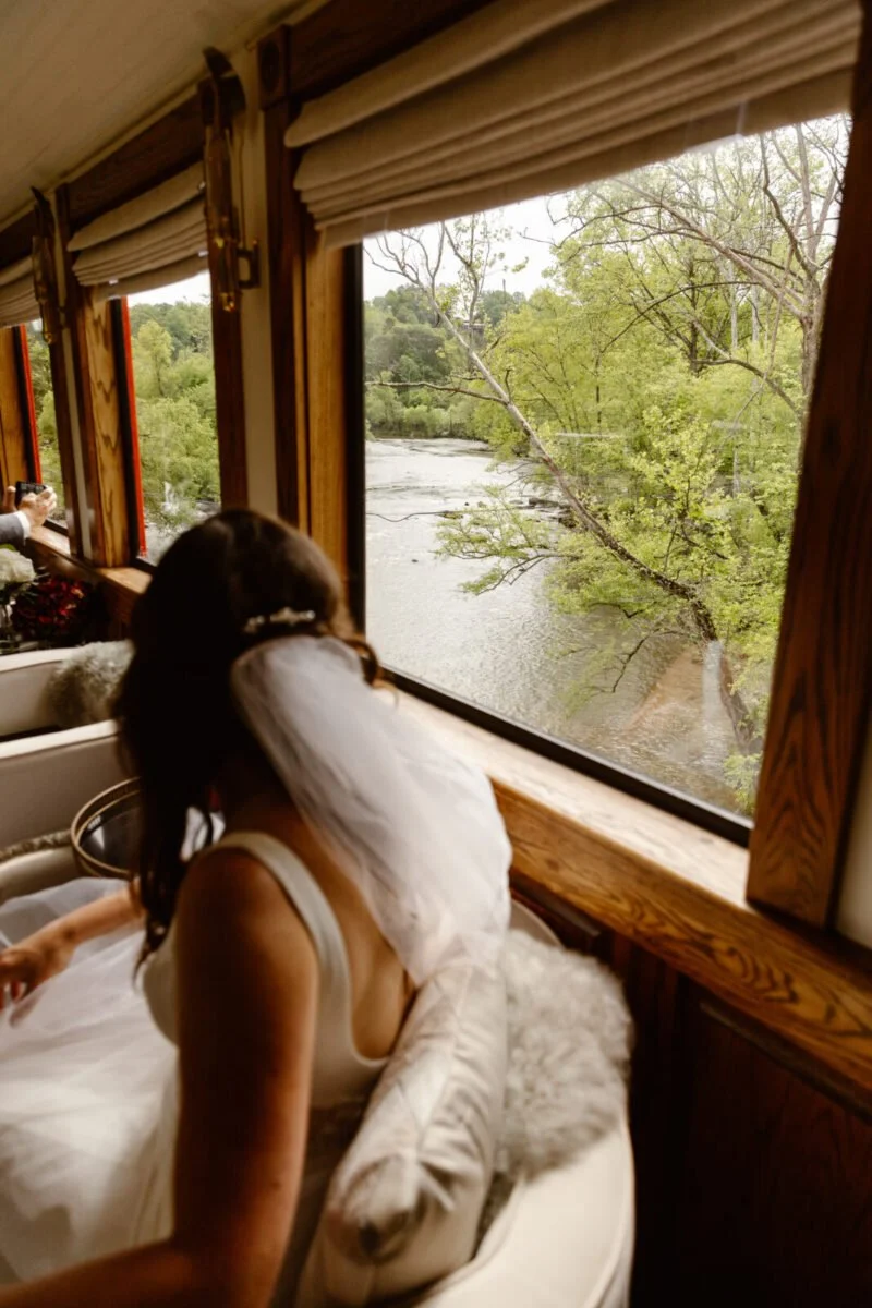 A woman with a veil and white dress sitting on a cushioned bench by a window overlooking a river and green trees.