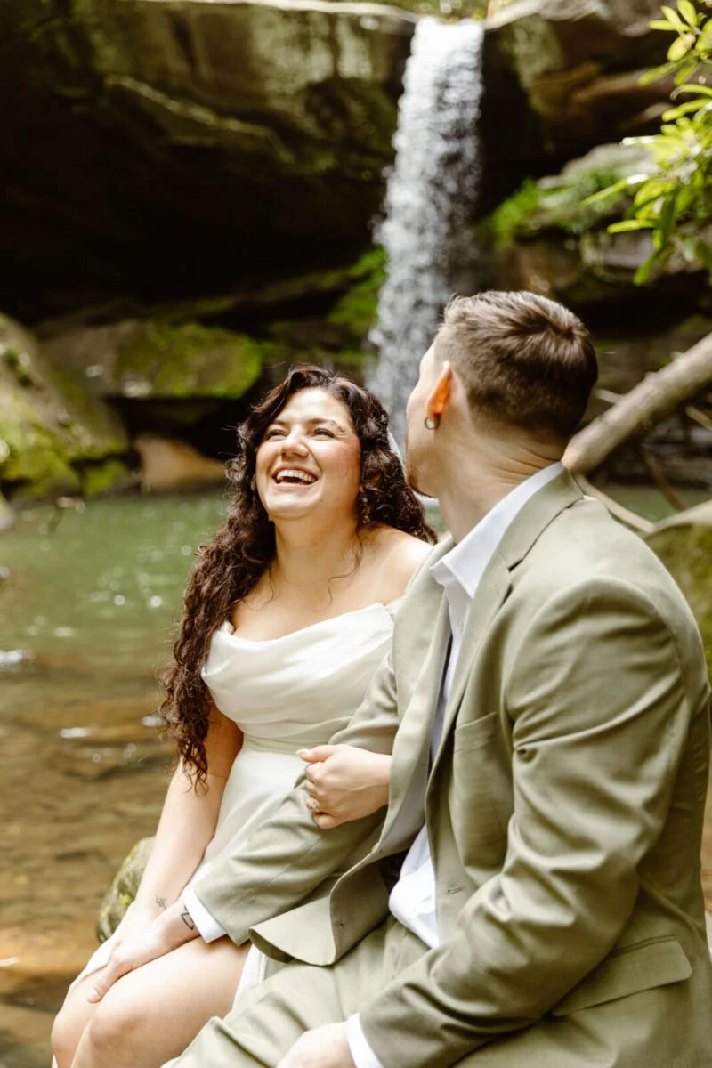A man and woman sitting by a river with a waterfall in the background, smiling at each other.