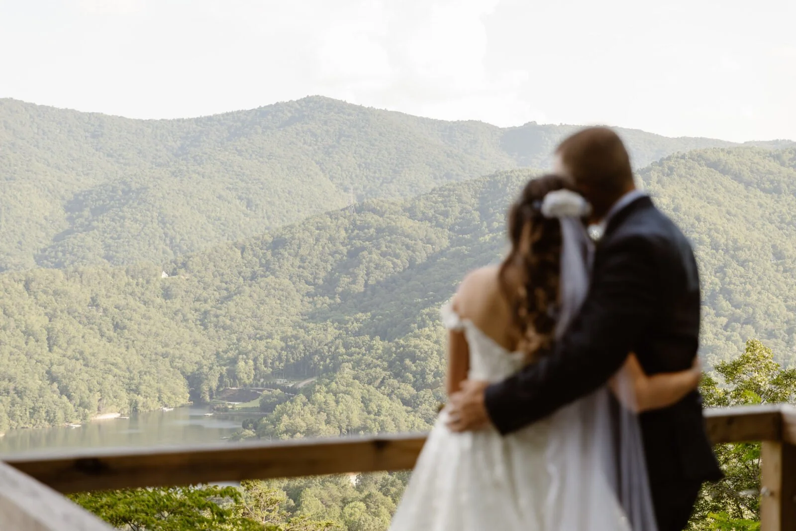 A couple dressed in wedding attire embracing with a scenic mountainous landscape and a river in the background.
