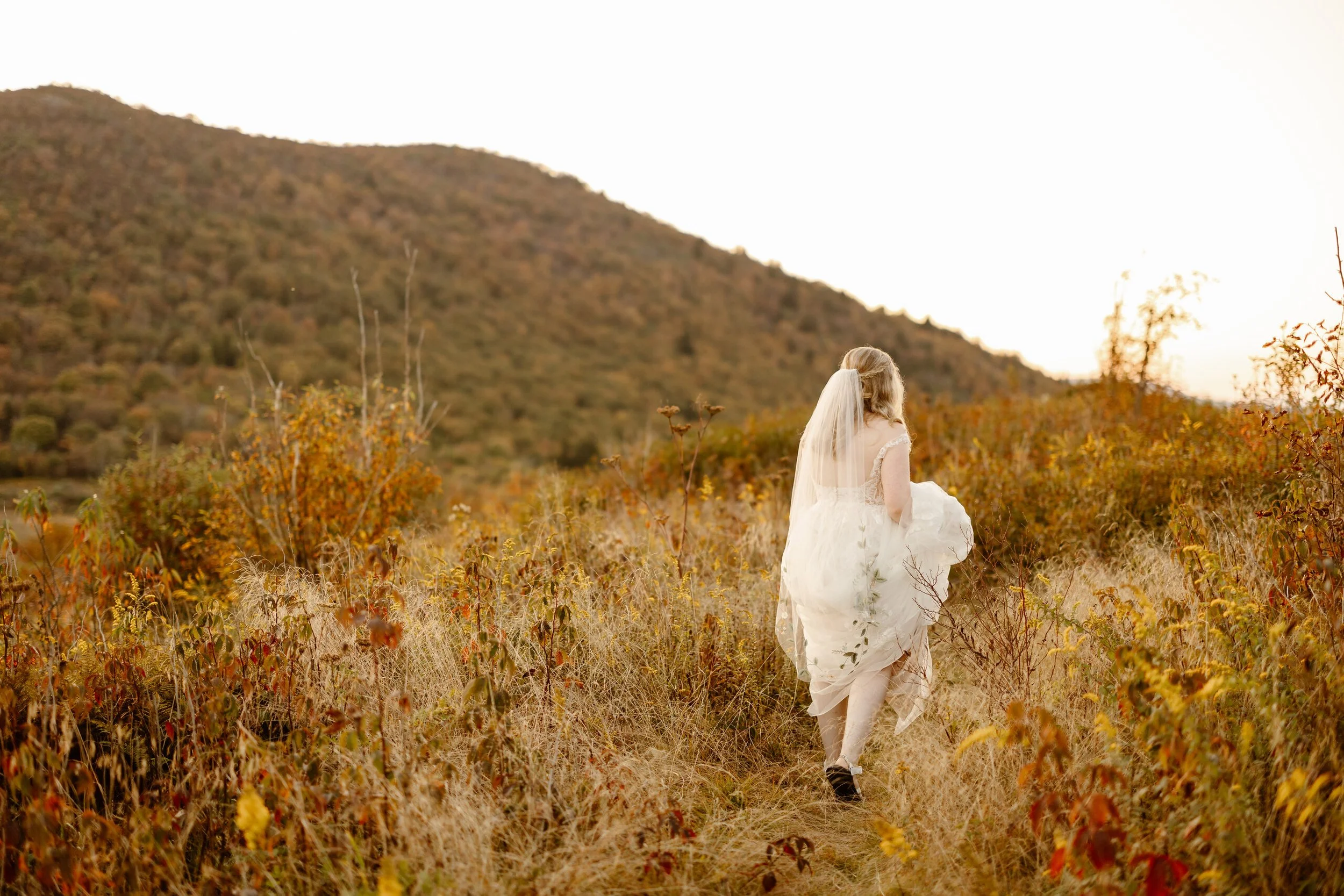 A woman dressed in a white gown and veil walking through a field of dry grass and autumn-colored bushes with a mountain in the background at sunset.