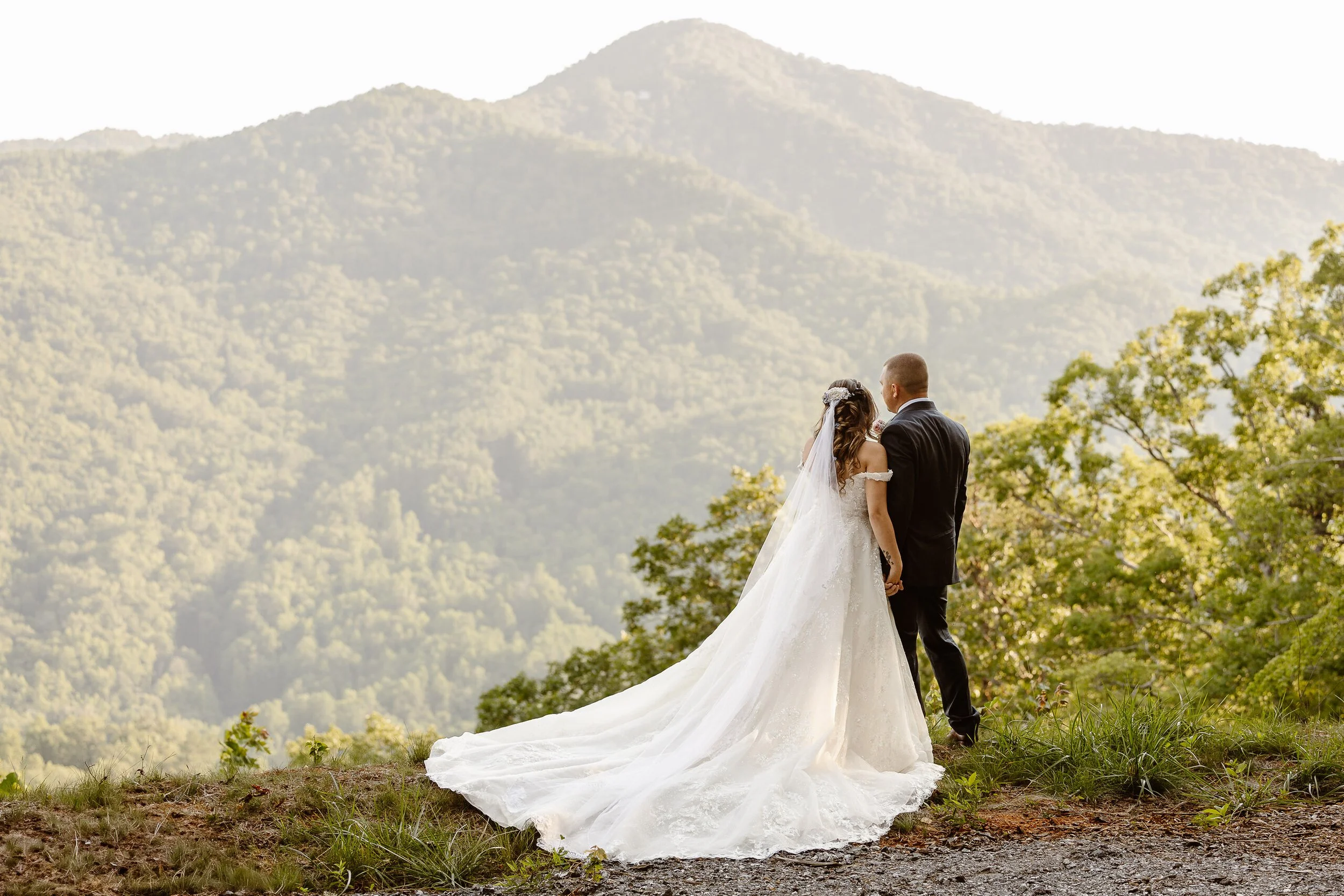Bride and groom standing together outdoors, overlooking mountains and trees, during a wedding ceremony or photo session.