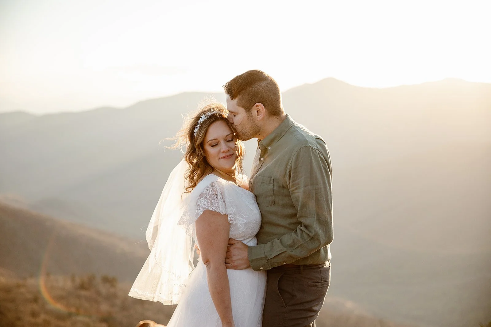 A bride and groom embracing outdoors during sunset, with mountainous landscape in the background.