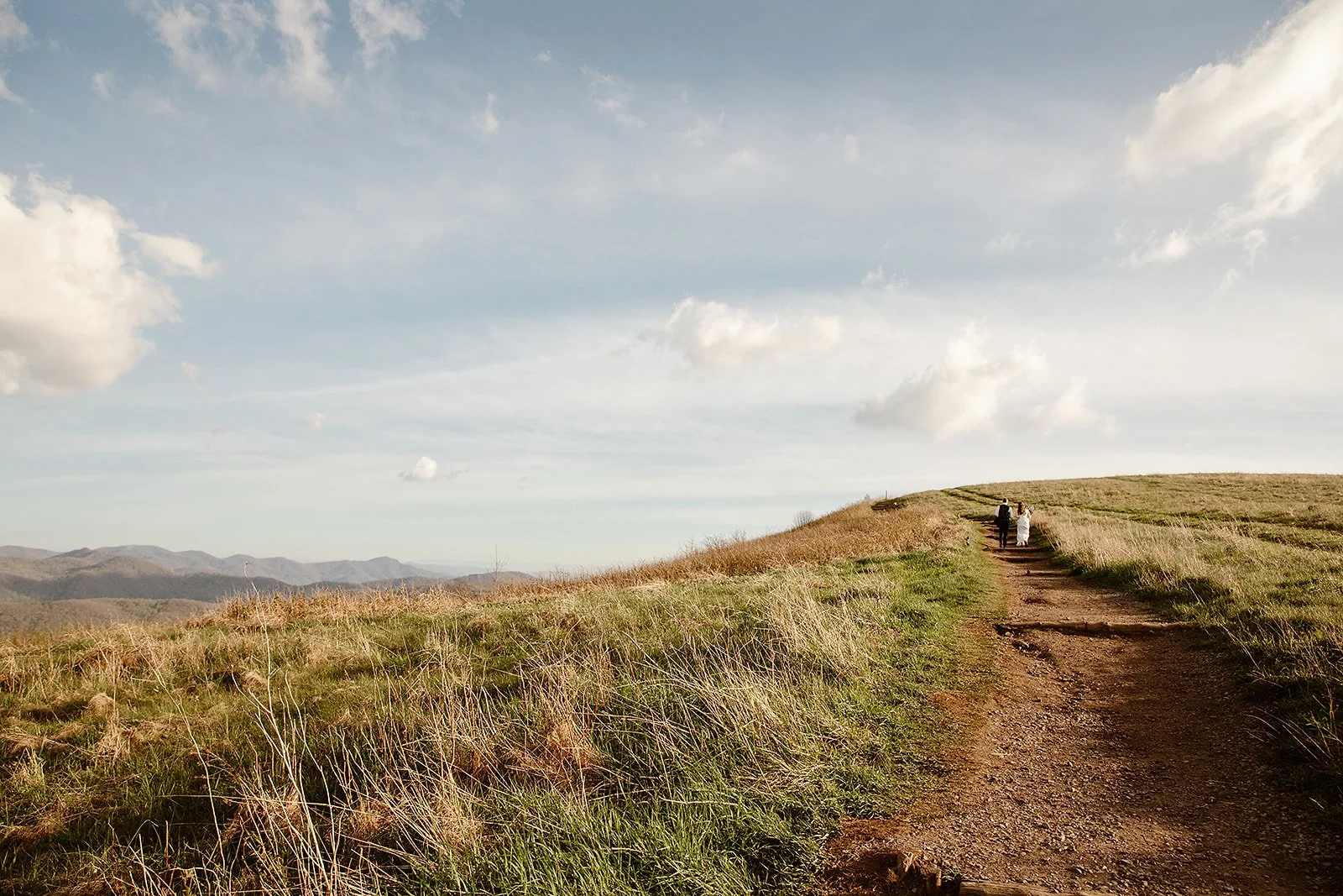 A dirt path through grassy hills leading up to a cloudy sky, with a couple walking along the trail.