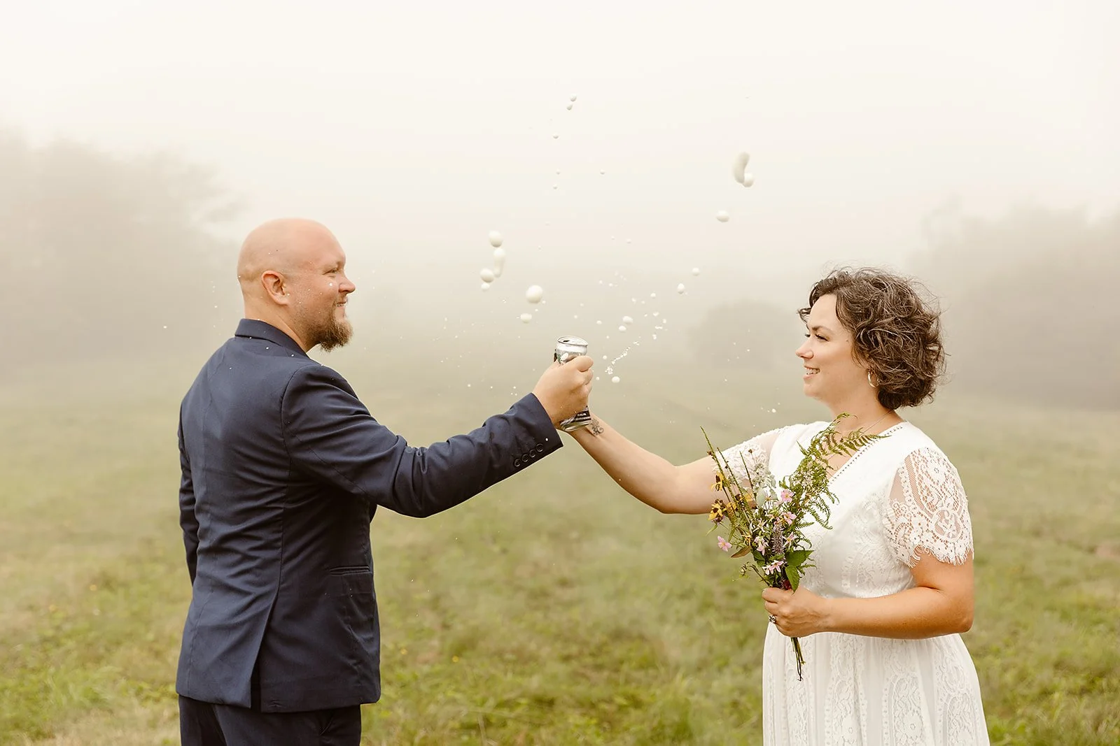 A man in a dark suit and a woman in a white lace dress celebrate with a champagne toast outdoors, with the woman holding a bouquet of flowers.