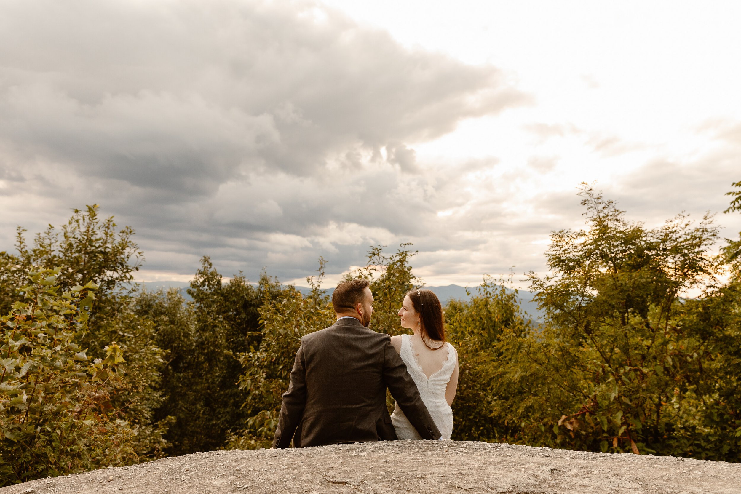 A couple sitting on a rock, looking at each other, with trees and mountains in the background and cloudy sky above.