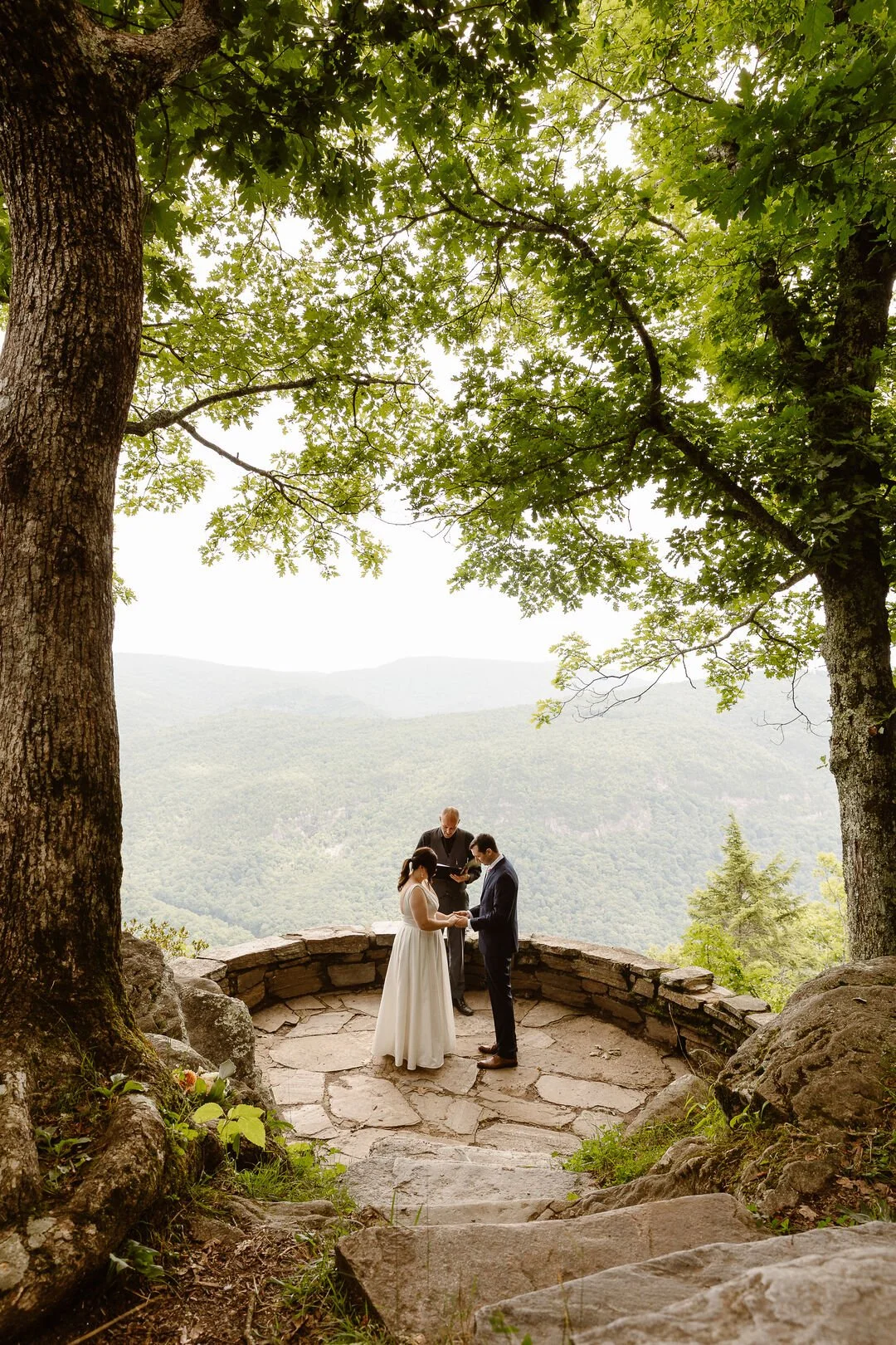 A couple getting married outdoors on a stone platform under large trees, with a scenic mountain view in the background. The officiant is standing behind them.