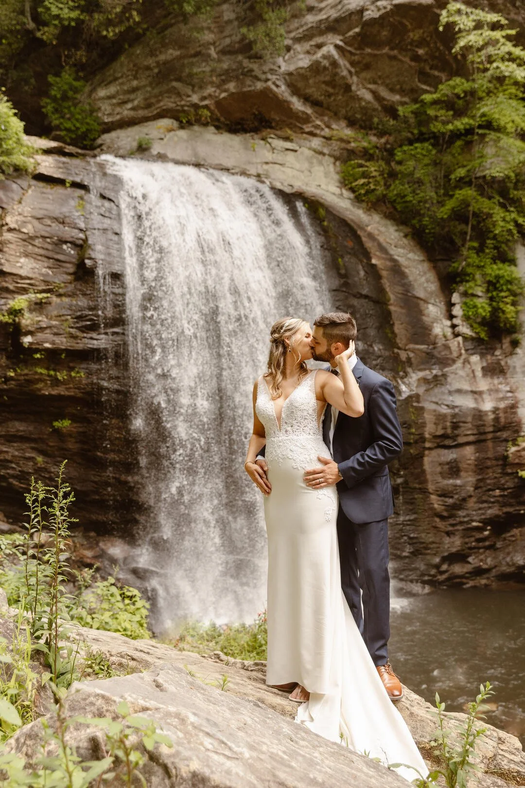 A bride and groom kissing in front of a waterfall in a natural outdoor setting.