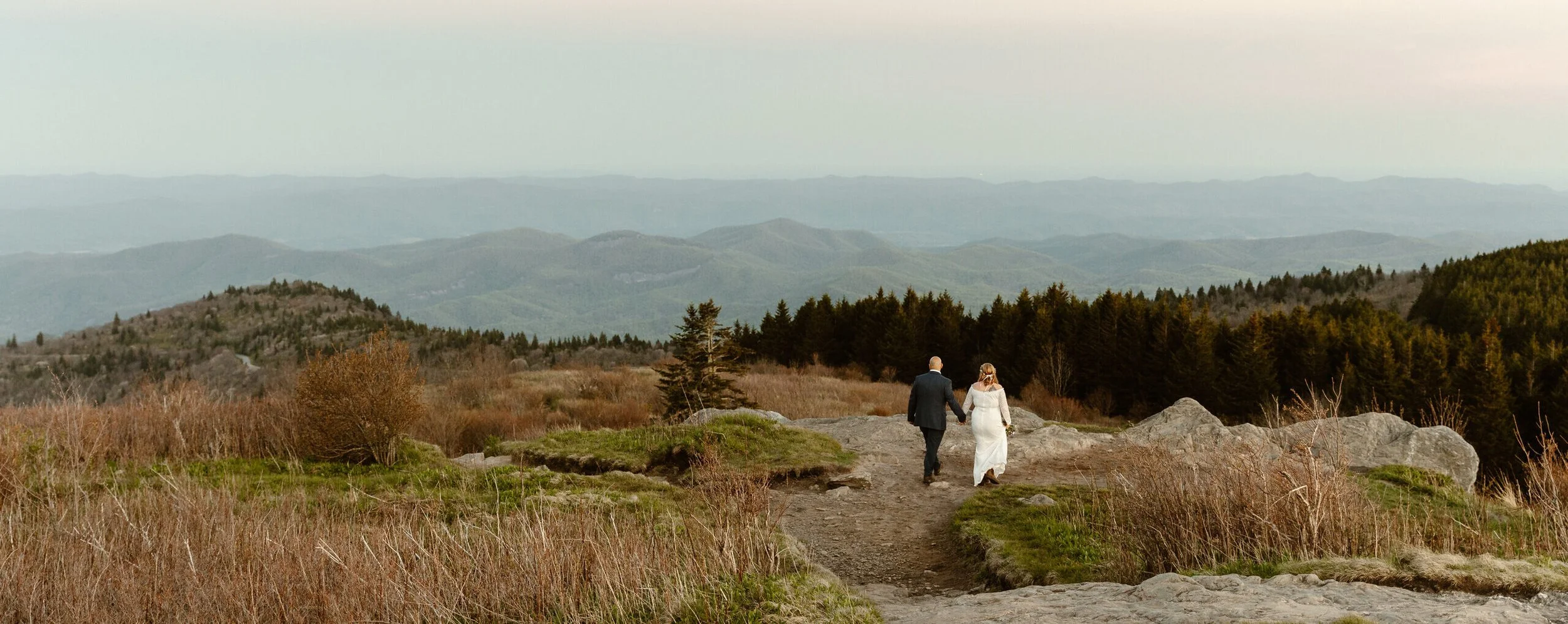 A bride and groom walking hand in hand on a rocky trail in a mountain landscape during sunset.