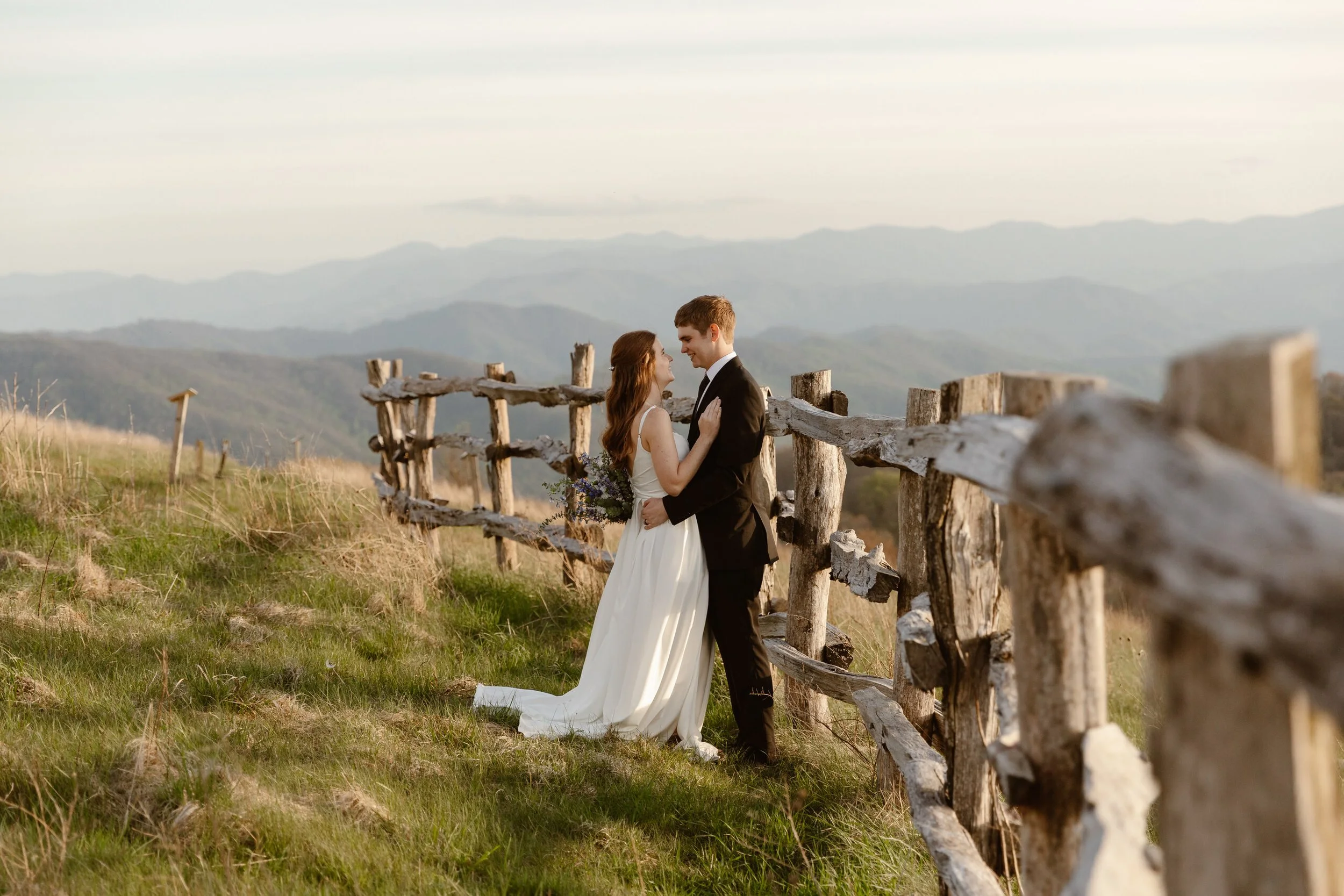 A bride and groom embrace on a grassy hillside with a wooden fence and mountain landscape in the background during sunset.