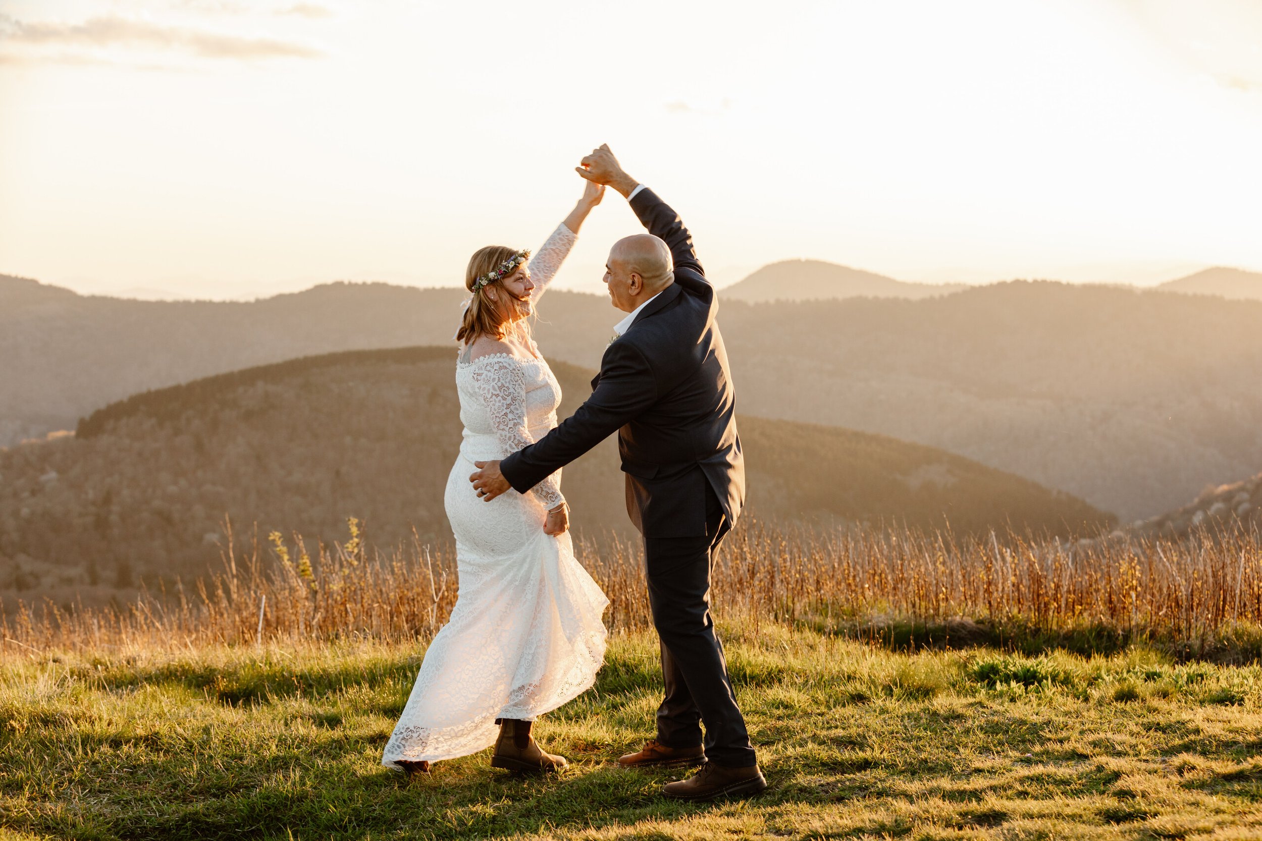 A newlywed couple dancing outdoors in a field during sunset, with hills in the background. The bride wears a white lace dress and floral headpiece, while the groom is in a dark suit. They hold hands and smile at each other.
