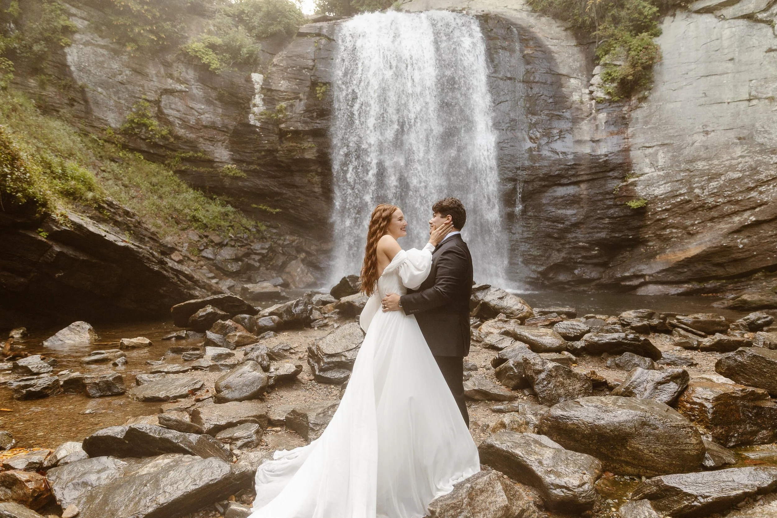 A bride and groom standing on rocks in front of a waterfall, embracing each other during a wedding photoshoot.