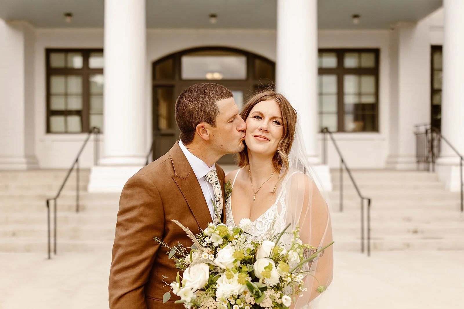 A groom kissing a bride on the cheek in front of a courthouse or government building, with steps and large white columns, during a wedding.