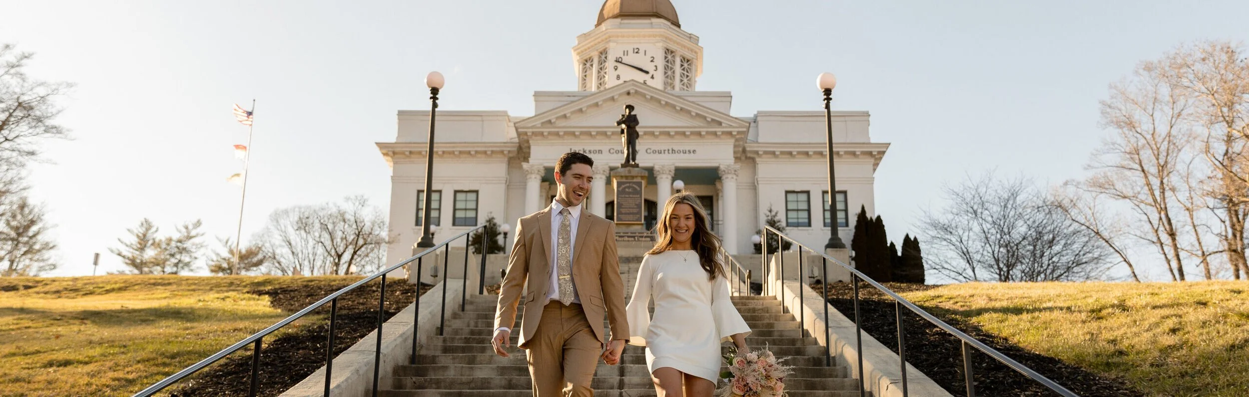 A smiling couple holding hands and walking down the stairs in front of a courthouse, with a woman holding a bouquet of flowers, during daytime with clear skies and leafless trees in the background.