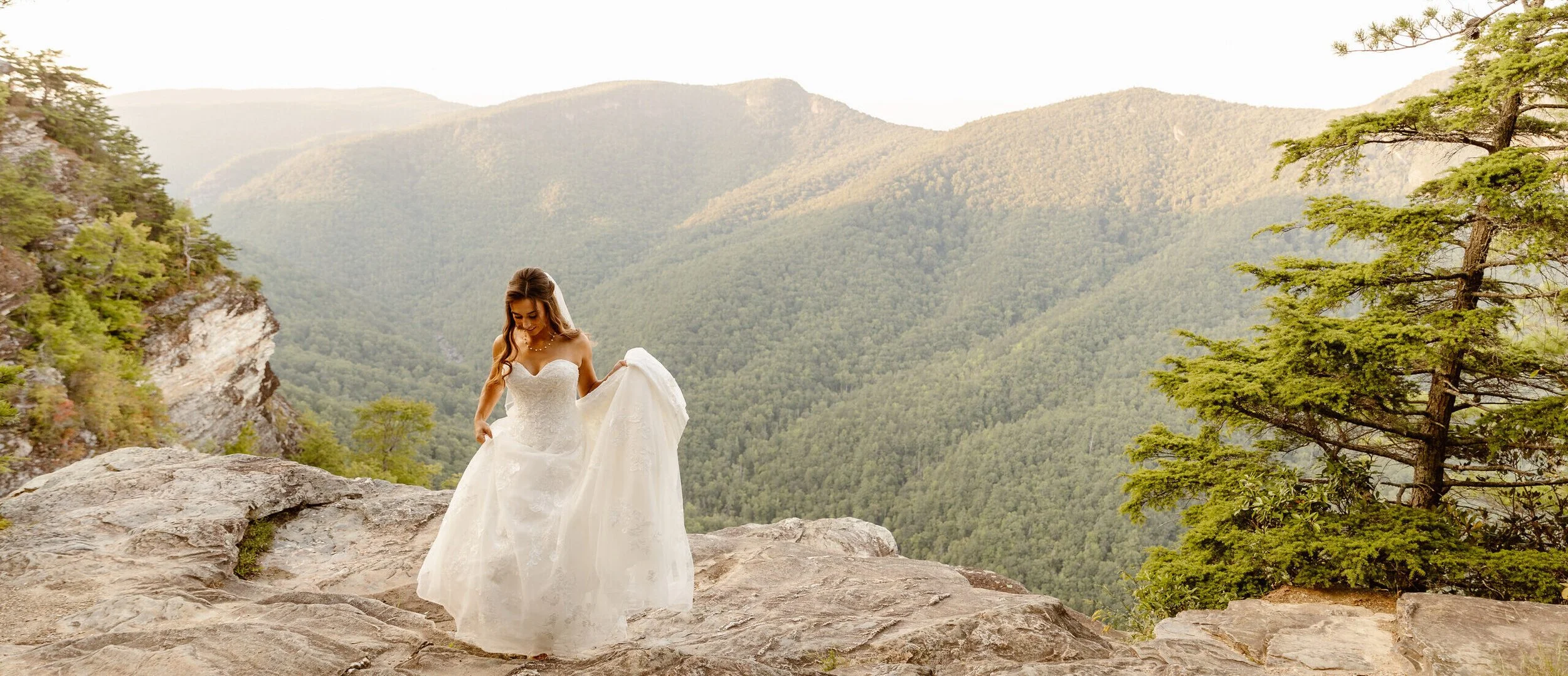 A woman in a white wedding dress standing on rocky terrain with a mountain landscape and trees in the background.