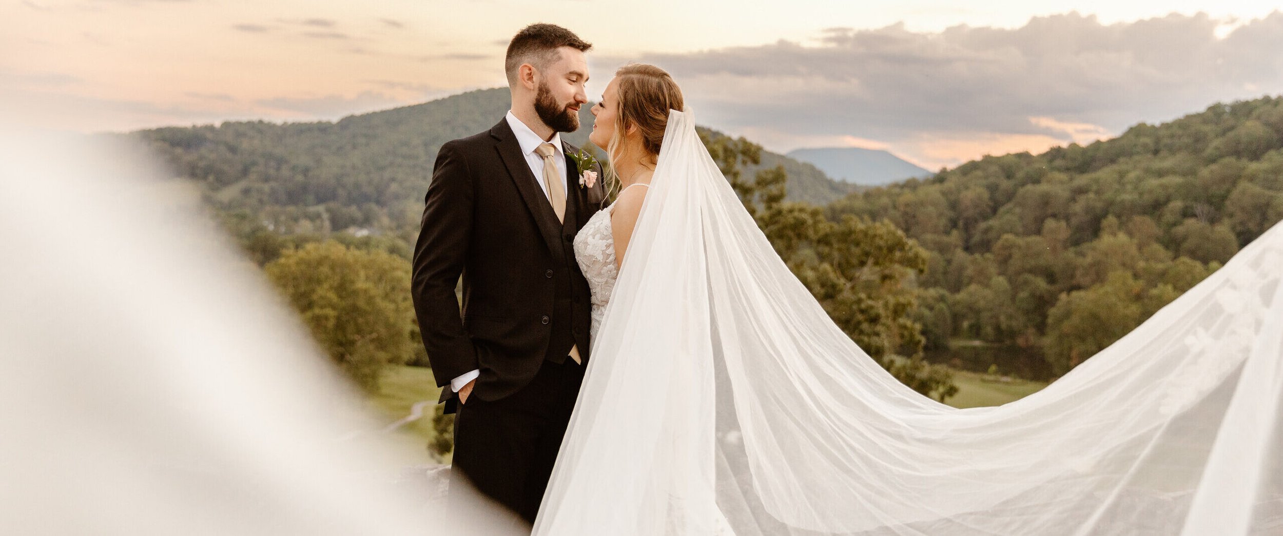 Bride and groom standing close, gazing at each other, outdoors during sunset with mountains and trees in the background.