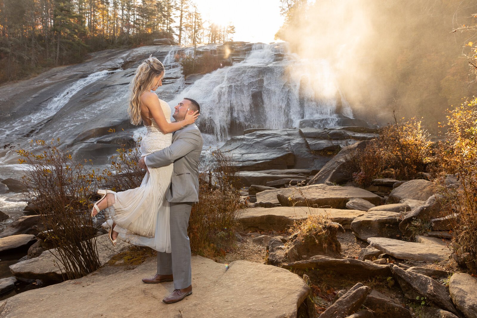 A man in a gray suit lifting a woman in a white wedding dress in front of a waterfall in a natural outdoor setting at sunset.